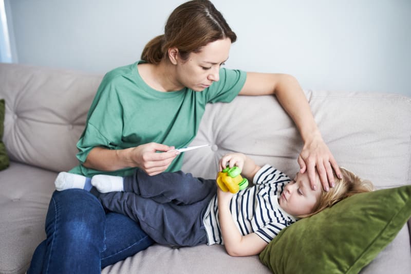 Mother taking young son's temprature while he lays on couch. 