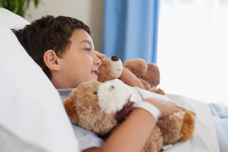 Young boy laying in hospital bed holding teddy bear.