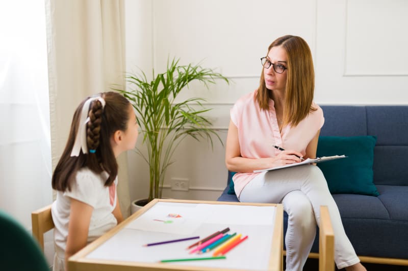 Young girl drawing art therapy session while counselor observes.