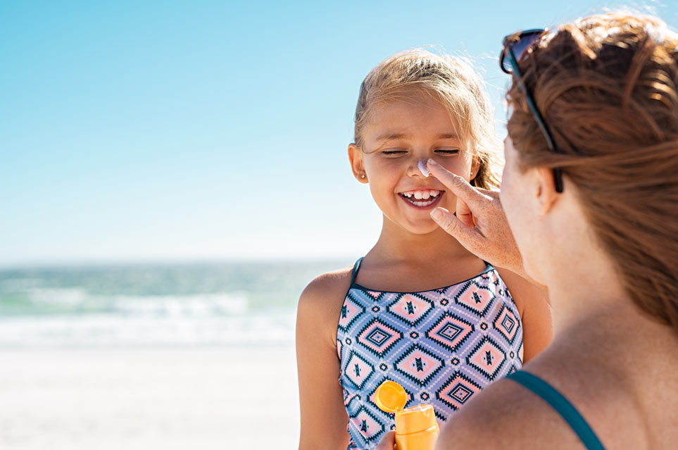 Young mother applying protective sunscreen on daughter nose at beach. 