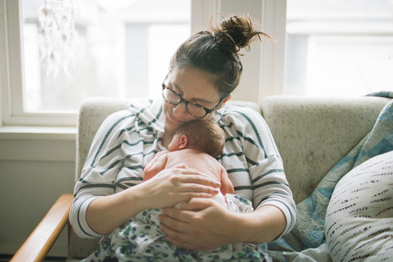 A mother holds her newborn baby close to her chest while seated on her sofa at home.