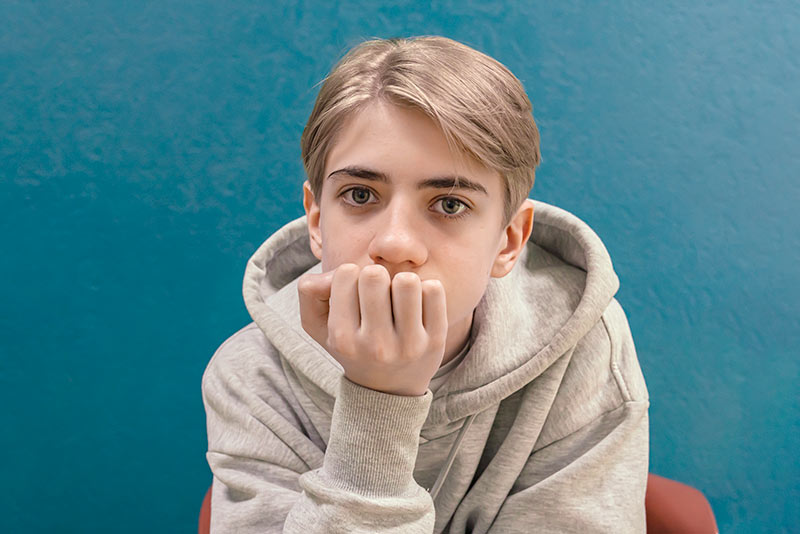 Portrait of a teenage boy, in a hoodie on a blue background, pensively looking at the camera.