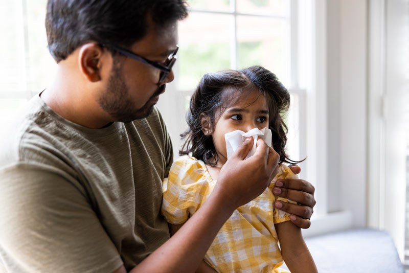 Father holding tissue to toddler daughter's nose.
