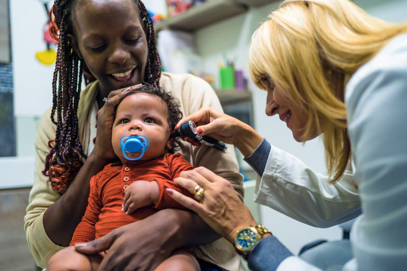 Baby sitting in mother's lap while doctor exams his ear with otoscope. 