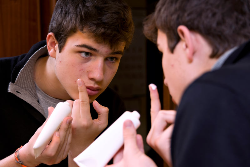 Teenage boy applying acne treament to face while looking in mirror.