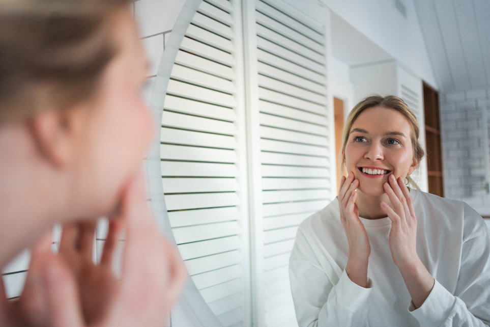 Happy young woman examines her face in mirror, satisfied with her skin youthful and healthy look.