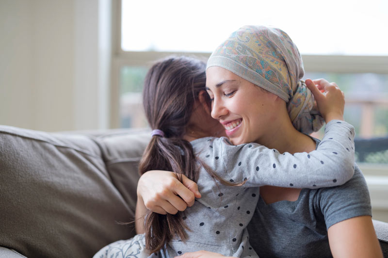 An ethnic young adult with cancer is hugging her child and smiling.