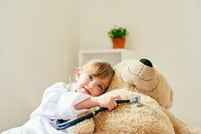 Adorable little girl dressed up as a doctor and examining a teddy bear with a stethoscope.