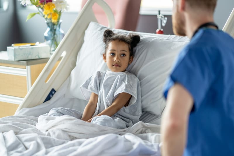 Young girl laying in hospital bed recovering from surgery as male nurse checks on her. 