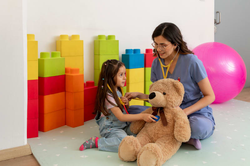 Young girl pretends to listen to teddy bear's heart during play therapy session.