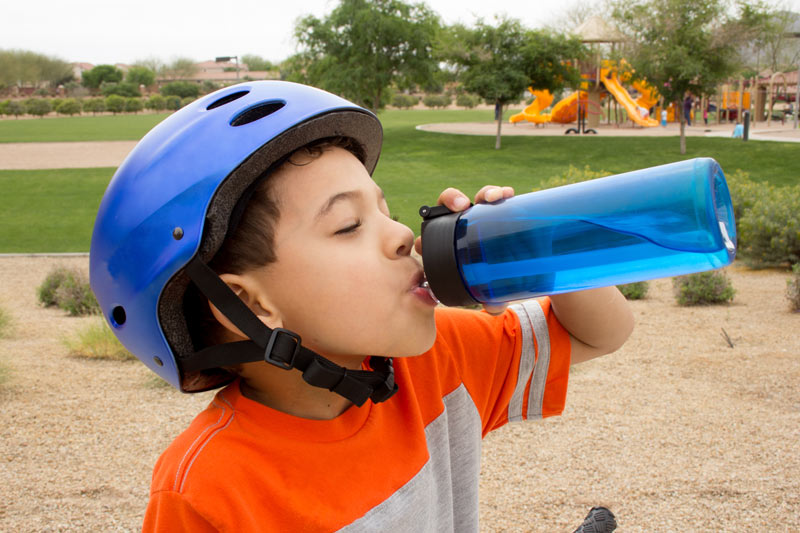 Thirsty boy drinking water outdoors wearing a bicycle helmet.